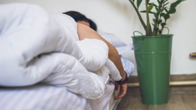 Person lying in bed with arm slung over the side of the bed. White duvet is pulled up, we cannot see their face. Green metal bucket vase sits to the right with dried foliage